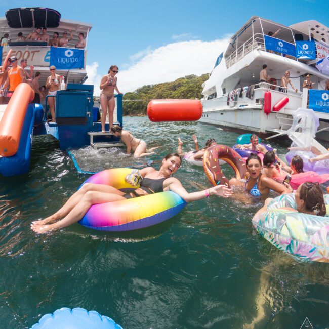 People enjoying a sunny day on the water with colorful inflatables. Boats are docked nearby with more people relaxing and socializing. The sky is clear with occasional clouds, and the water is calm, creating a lively and cheerful atmosphere.