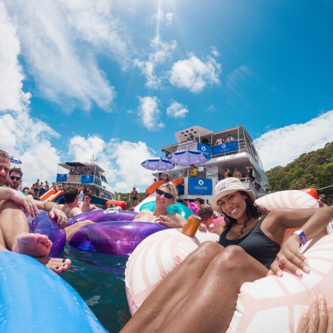 A group of people relax on colorful inflatable floats in the water near a boat, enjoying a sunny day. The sky is bright with a few clouds. Two main figures are smiling, one holding a drink. Large boats are anchored in the background.