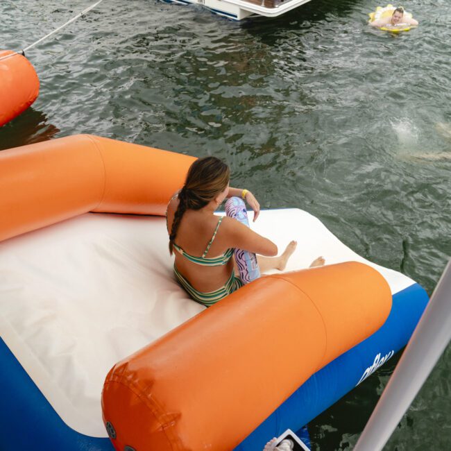 A person sits on an orange and blue inflatable raft on the water, holding onto another inflatable ring. Other people are seen swimming nearby. There is a boat visible in the background.