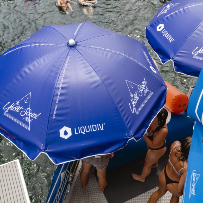 People enjoying a pool party on a yacht, with blue umbrellas labeled "Liquid IV" providing shade. Swimmers in the water float on colorful inflatables, while others stand on deck in swimwear.