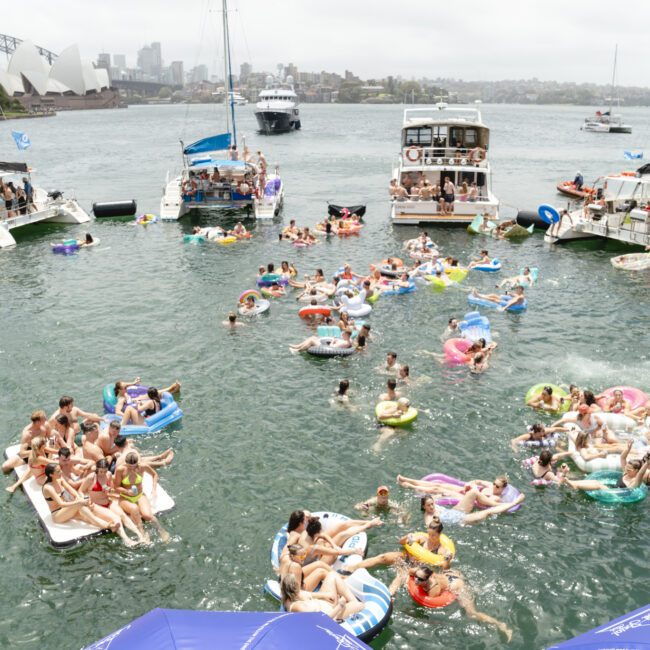 A lively scene on the water features groups of people on inflatable floats and boats, enjoying a sunny day. The Sydney Harbour Bridge is visible in the background, adding to the vibrant atmosphere of the gathering.