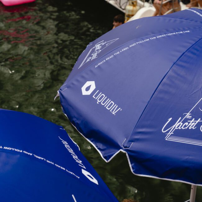 Blue umbrellas with "The Yacht Social Club" and "LIQUIDIV" logos are open near a body of water. Partially visible people and floating inflatables are in the background.