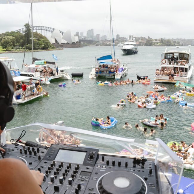 DJ at a mixing console on a boat, overlooking a party scene on the water with numerous people on inflatable rafts and boats. The Sydney Opera House and Harbour Bridge are visible in the background under a cloudy sky.