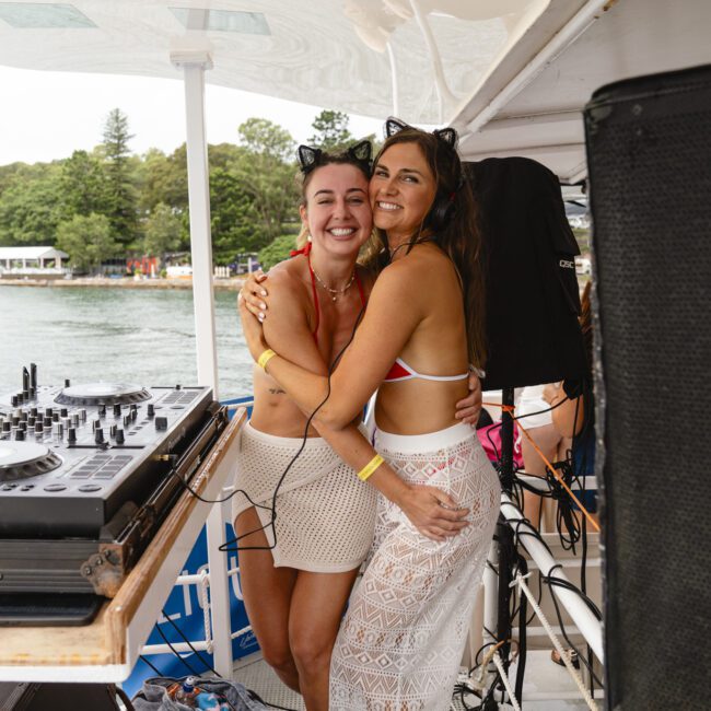 Two smiling women on a boat pose beside DJ equipment. One wears a red bikini top and white shorts, the other wears a black bikini top and long lace skirt. They are surrounded by a scenic view of water and greenery.