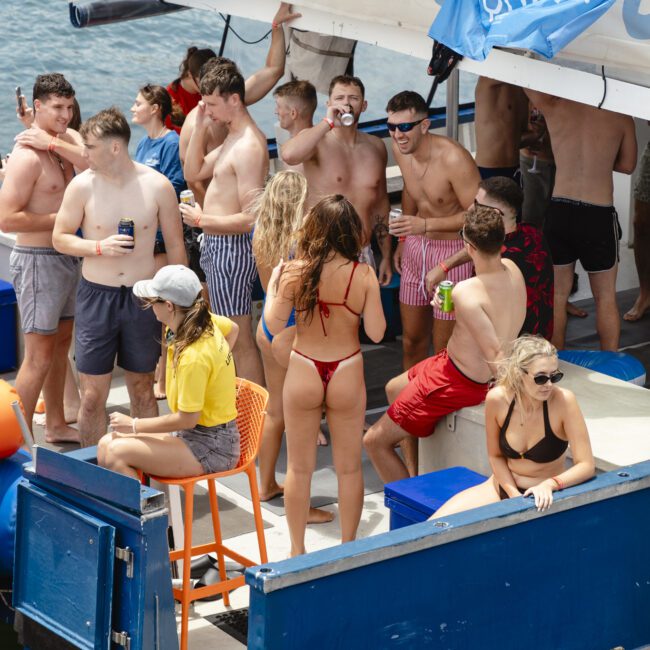 A group of people in swimwear are socializing on a boat under a shaded area. Some are sitting, others standing, talking, and holding drinks. The boat is decorated with banners, and it's a sunny day.