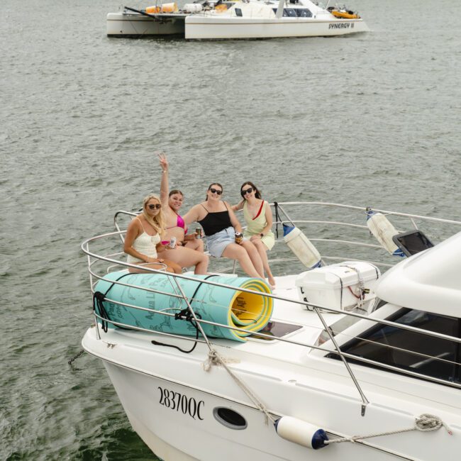 Four people sitting and relaxing on the deck of a white yacht. They are wearing swimsuits and sunglasses, with one person waving. Two large floats are rolled up next to them. In the background, another boat on the water.