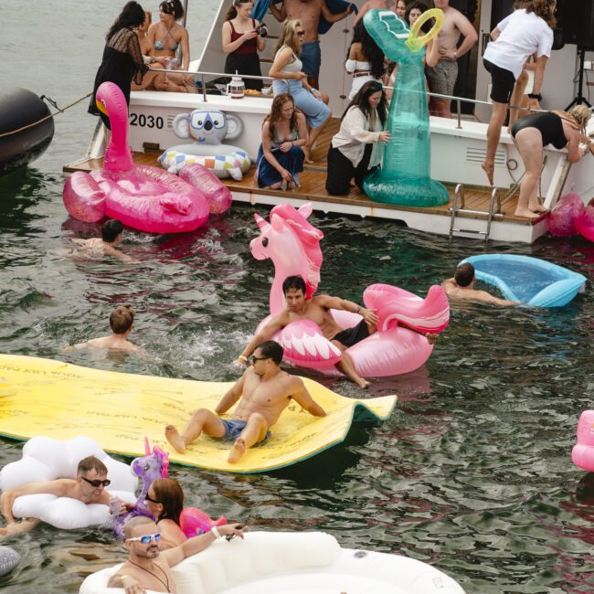 People enjoy a boat party, floating in a lake with colorful inflatables including a unicorn, flamingo, pizza slice, and lounge chair. The docked boat is filled with more people, some in costume, as music plays.