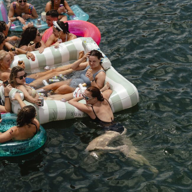 A group of people are relaxing in a lake on various inflatable floats, including striped and donut-shaped ones. They are in swimsuits, enjoying a sunny day on the water.