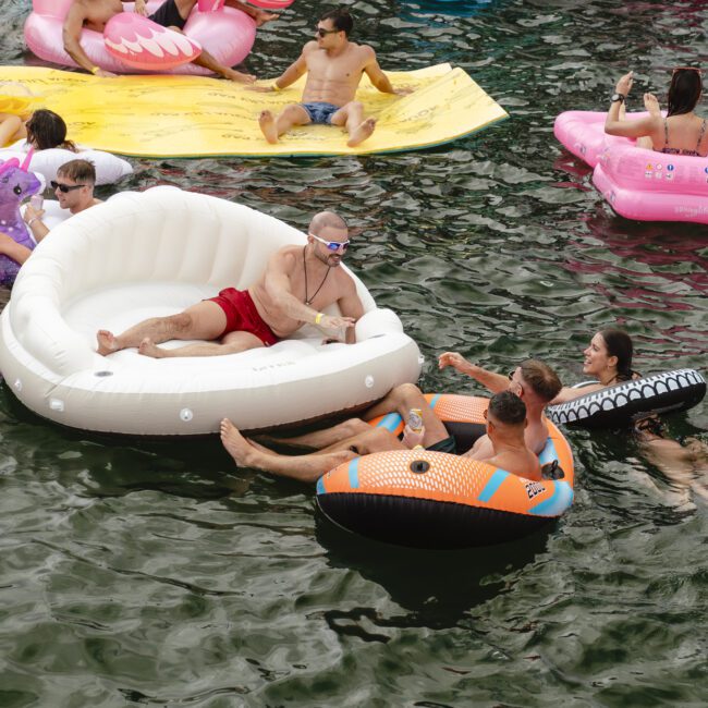 A group of people relaxing on inflatable floats in a body of water. Floats include a white couch, a black and orange inner tube, and a pink flamingo. Everyone is enjoying the sunny day.