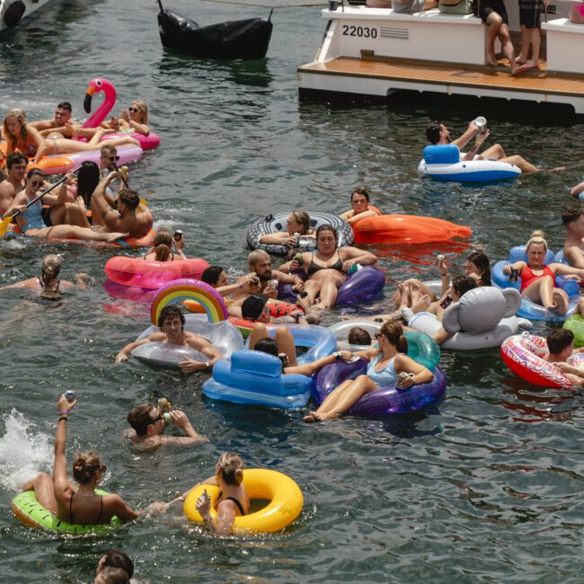 People enjoying a sunny day on the water, floating on colorful inflatable tubes. A boat docked nearby has more people. The scene is lively, with pink flamingo, rainbow, and animal-shaped inflatables among the crowd.