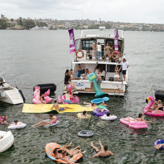 A lively boat party with people enjoying the water on colorful inflatables. Several individuals are on a large boat, while others float nearby on swans, donut rings, and other shapes. The backdrop features a coastal landscape under a cloudy sky.