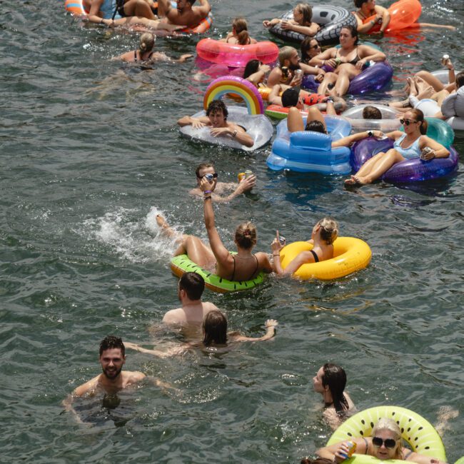 A crowd of people floats on colorful inflatable tubes in a body of water. They are enjoying a sunny day, some holding drinks and others chatting. The inflatables vary in shapes, including donuts and flamingos.