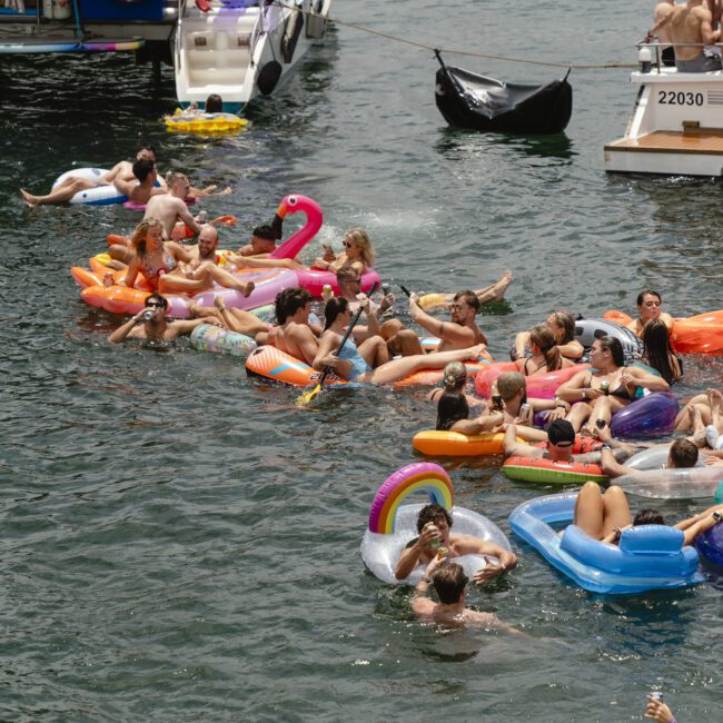 People float on colorful inflatables, like a flamingo and unicorn, in a sunny lakeside scene. Boats are nearby, and the water is crowded with cheerful swimmers enjoying the day.