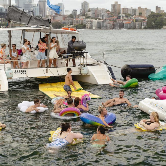 People enjoying a boat party on the water, with several individuals floating on inflatable toys like flamingos and unicorns. The backdrop includes city buildings and another boat. The atmosphere is lively and festive.