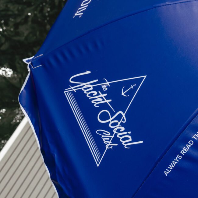 Close-up of a blue umbrella with "The Yacht Social Club" logo and the text "Always read the label and follow the..." in white. Part of the wooden dock and water can be seen in the background.