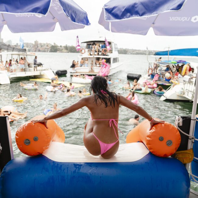 A woman in a pink bikini slides down an inflatable slide into a body of water, surrounded by people on boats and inflatables. A lifeguard in a yellow shirt observes nearby. Umbrellas provide shade, and the scene looks lively and summery.