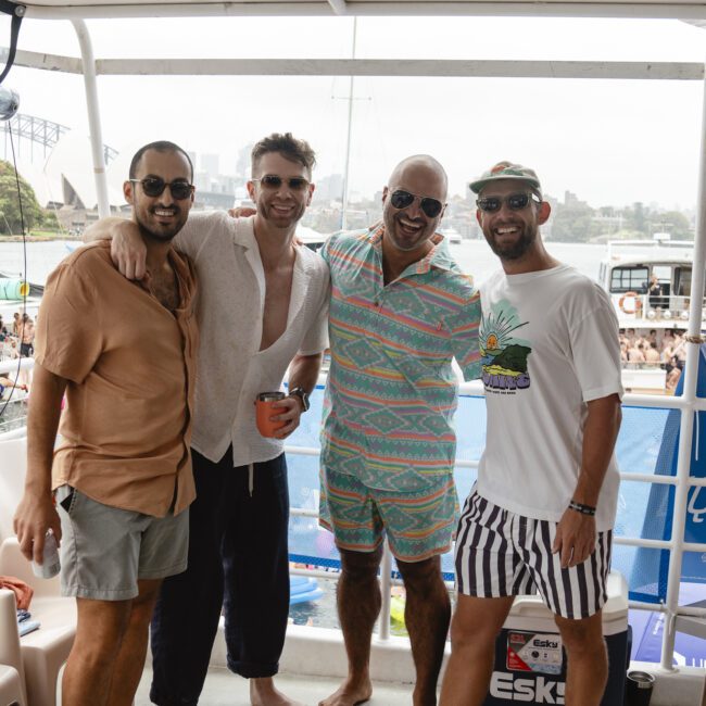 Four men smiling and posing together on a boat. They are wearing casual summer clothing. The ocean and other boats are visible in the background, along with the Sydney Opera House and Harbour Bridge.