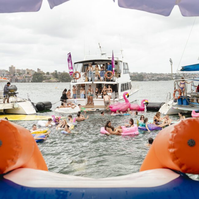 A lively group of people enjoy a party on the water, floating on inflatable toys near anchored boats. The scene is festive with colorful inflatables including a large pink flamingo. In the foreground, an inflatable orange barrier frames the view.