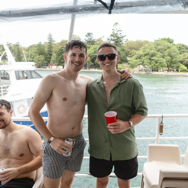 Two men are smiling on a boat. One is shirtless wearing striped shorts, the other is in a green shirt and dark shorts, holding a red cup. They stand near a seated person. The background features trees and water.