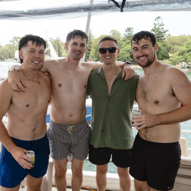 Four men in swimwear and casual clothes stand together on a boat, smiling at the camera. Each holds a beverage can. The background shows water and trees under a cloudy sky.