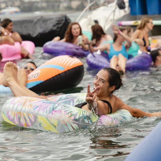 A woman smiling and giving a peace sign while floating on an inflatable tube in a body of water. Surrounding her are people lounging on other colorful inflatables, enjoying the water under a cloudy sky with boats in the background.