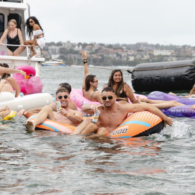 People enjoying a sunny day on the water, floating on inflatable tubes and loungers. Some are lounging on a boat, while others are paddling in the water, laughing and wearing swimwear. Background shows a distant shoreline with buildings.