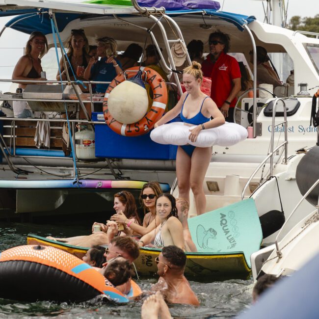 A group of people enjoys a sunny day on the water near two boats. Some are in the water on inflatable floats, while others lounge on the boats. One person, wearing a blue swimsuit and inflatable ring, stands on a boat. Smiles and laughter abound.