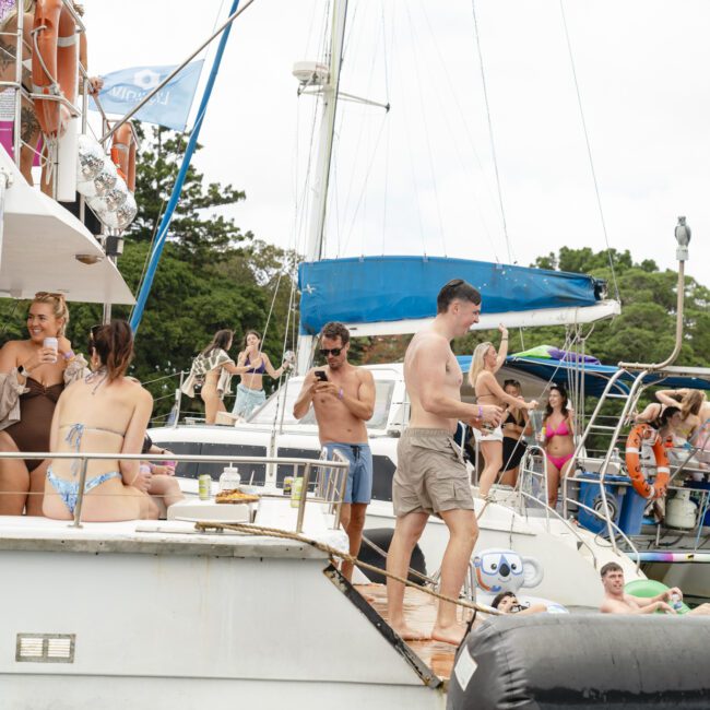 A group of people on a boat, enjoying a sunny day. Some are sitting and chatting, others are standing or walking around. Another boat is docked nearby with more people on it. The surrounding area has greenery and trees.