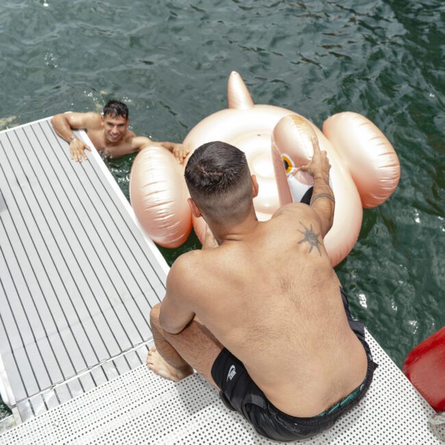 A man sits on a white dock with his feet in the water, reaching for a large, inflatable pool float shaped like a peach and pink bear. Two people swim nearby with colorful floats, surrounded by green water.