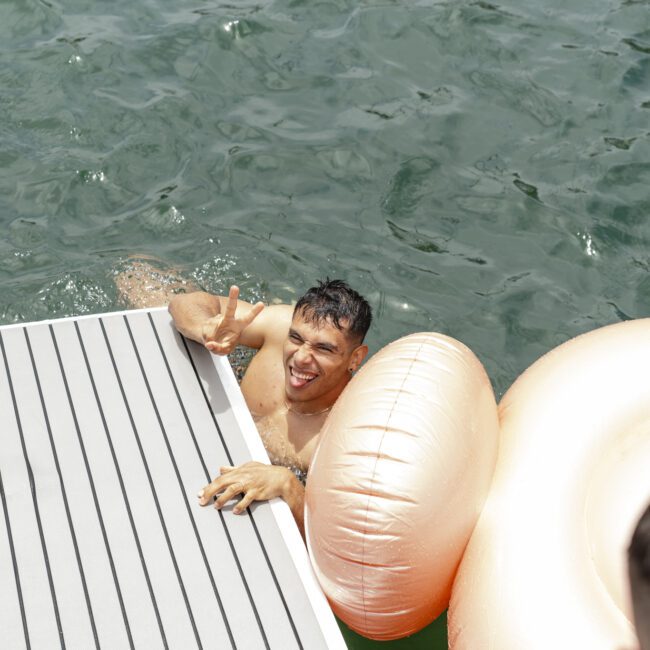 A man in a lake smiles and flashes a peace sign while holding onto a floating dock. Beside him, there is a large, inflatable, light brown or gold float. The water is a deep green, and part of another person is visible in the foreground.