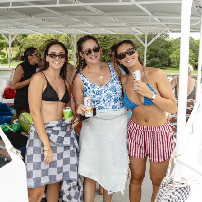 Three smiling women in swimwear stand on a boat deck holding beverages. The woman on the left is in a black bikini, the middle woman in a blue and white top, and the right woman in a blue top with red striped shorts. Trees are visible in the background.