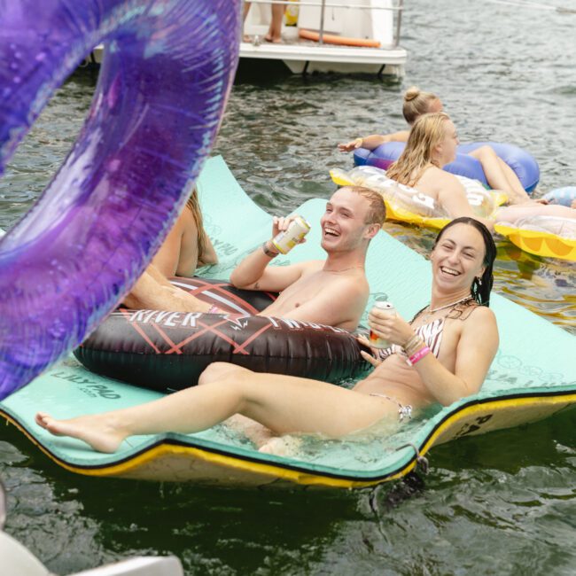 A group of people enjoying time in the water. Two people are smiling and relaxing on a foam mat, each holding a drink. Others float nearby on inflatable rafts. A boat is partly visible, and a purple inflatable is in the foreground.