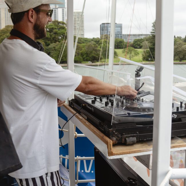 Man wearing a white shirt, cap, and striped shorts stands barefoot at a DJ deck on a boat. He's adjusting the mixer while overlooking a scenic background with green trees and tall buildings. The scene suggests a relaxed outdoor event.