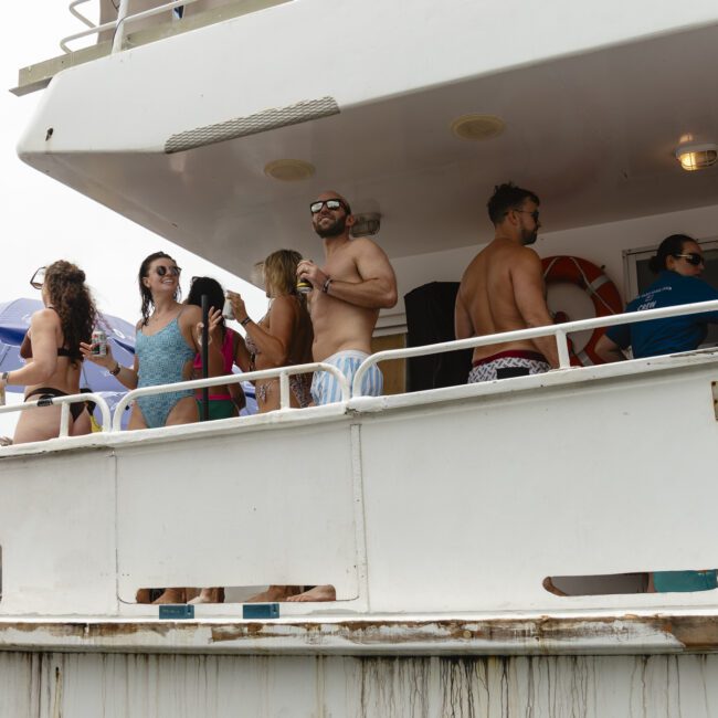 People are socializing on a boat deck, holding drinks and enjoying the view. Some are wearing swimsuits and sunglasses. There are umbrellas in the background, suggesting sunny weather. A banner labeled "Liquido" is partially visible.