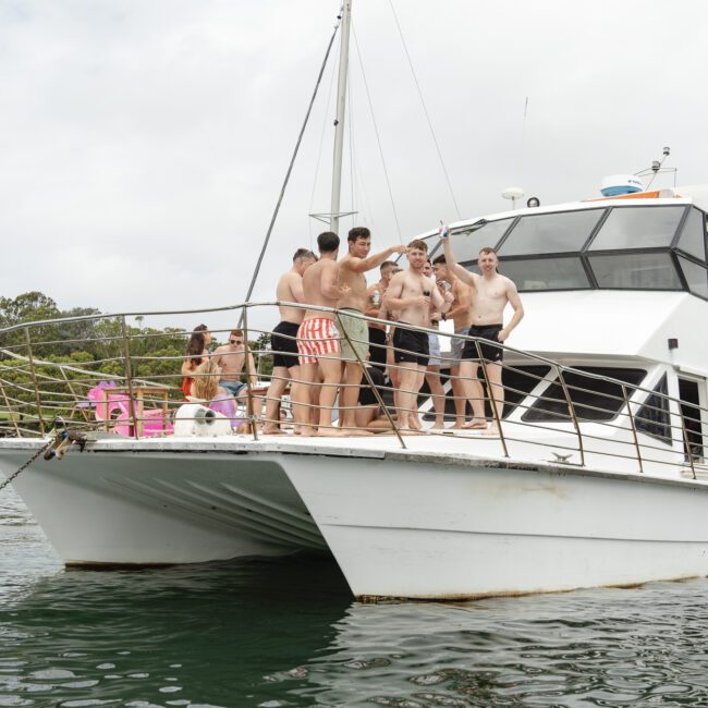 A group of people in swimwear are gathered on the deck of a yacht named "Kondor," anchored near a lush, wooded shoreline. The weather is overcast.