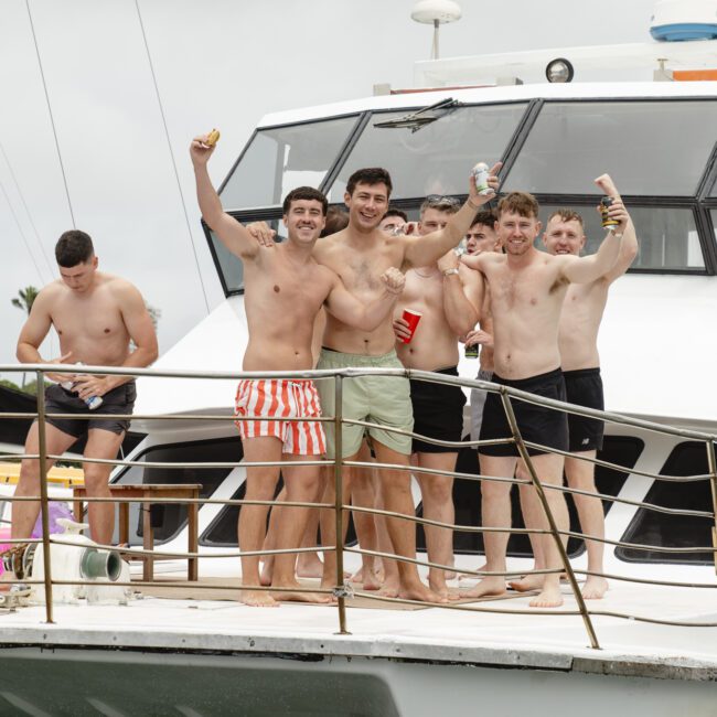 A group of six men in swimwear stand on the deck of a boat, smiling and holding drinks. Other people are visible in the background, enjoying the boat ride. The sky is overcast.