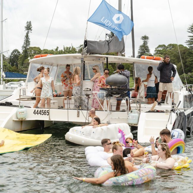 A group of people enjoying a sunny day on a catamaran and in the water. Some are lounging on inflatable floats, including a unicorn and a flamingo. The boat has a "Lidomare" flag and a grill at the back. Trees and other boats are in the background.