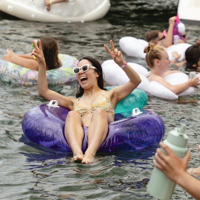 A woman in sunglasses smiles and raises her hands in a peace sign while floating on a purple inflatable in a lake. Around her, other people are relaxing on various floaties. The atmosphere is lively and joyful.