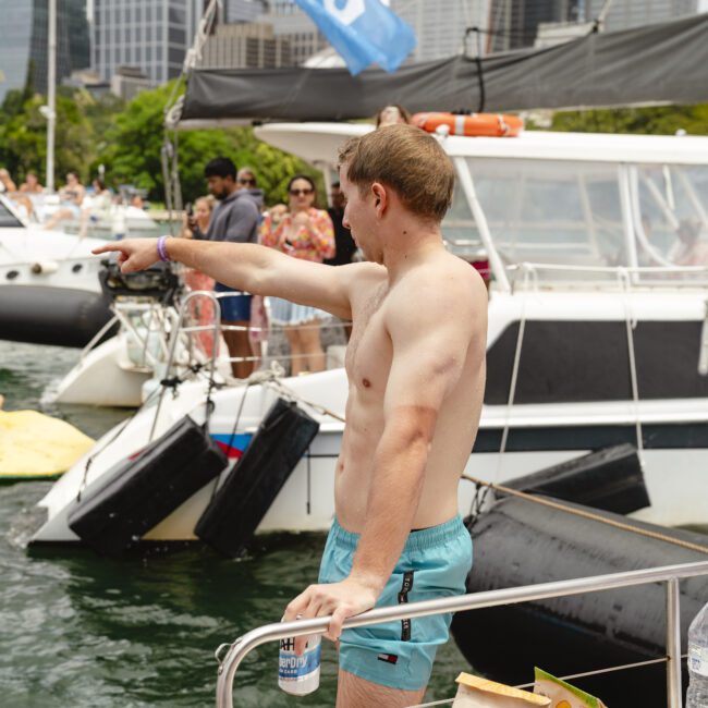 A shirtless man on a boat is pointing towards people lounging on a floating mat in the water. Other boats and people are nearby. Another person is using a smartphone. Buildings and greenery are visible in the background.