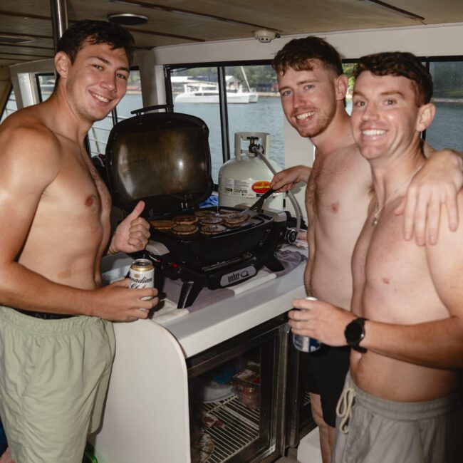 Three men stand inside a boat near a grill, smiling. They are shirtless and holding drinks, with one man giving a thumbs up. The background shows a window and a view of the water outside.