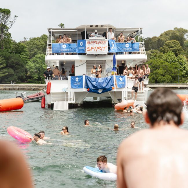 A large boat with "Barefoot Spirit" sign is moored on a sunny day, surrounded by people swimming and floating on inflatables in the water. Lush greenery is visible in the background. The scene is lively and festive.