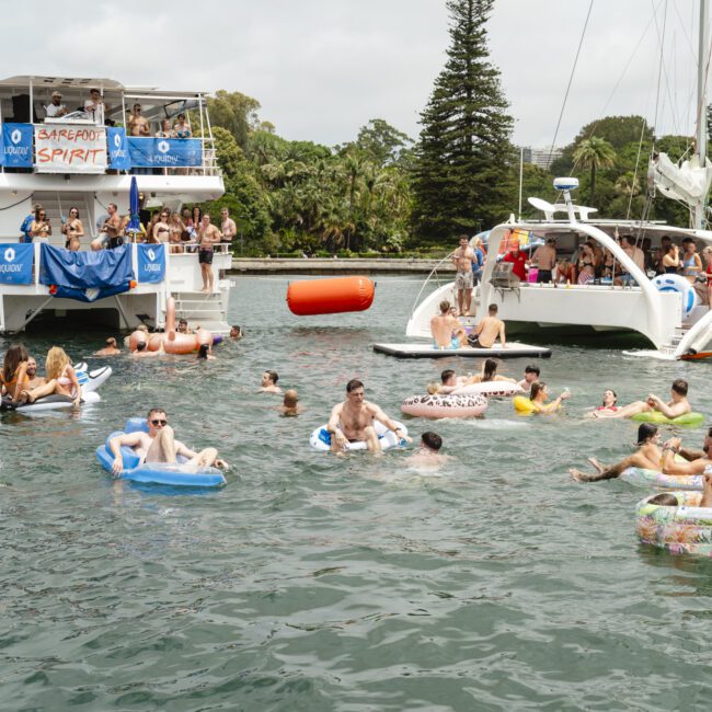 A lively group of people relaxing in a body of water on inflatable rafts and tubes. A double-decker boat with banners and a smaller sailboat are in the background. Trees and a few buildings are visible on the shore.