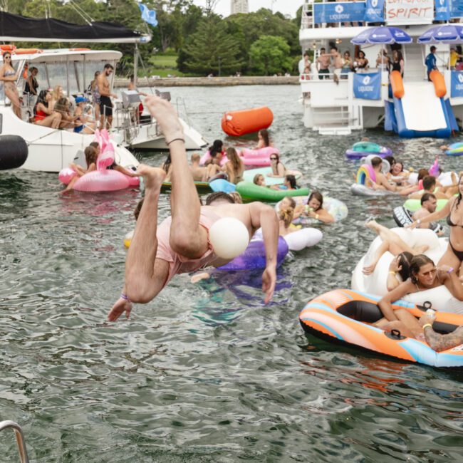 People enjoy a festive gathering on a lake with various inflatables. Some are diving into the water, while others relax on floaties. Boats are anchored nearby, and trees are visible in the background. The atmosphere is lively and cheerful.