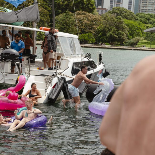 People enjoying a party on a boat, surrounded by colorful inflatable tubes. Some are in the water, and others are on the deck. The background features lush greenery and city buildings.