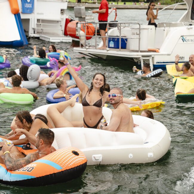 A lively scene on the water shows people enjoying a boat party. Individuals relax on inflatables, including a woman in a bikini posing joyfully with others around her. A boat is docked nearby, and an inflatable slide is visible.