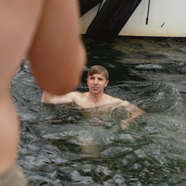 A shirtless man with short hair is in water near a docked boat, looking towards another person in the foreground. The boat has black fenders, and part of a logo that reads "Yacht Social" is seen in the corner.