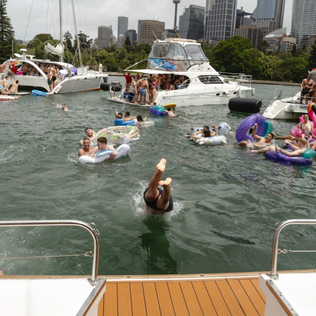 People are enjoying a sunny day on a lake, floating on colorful inflatables and swimming. Boats are docked nearby, and a person is diving into the water. The city skyline and trees are visible in the background.