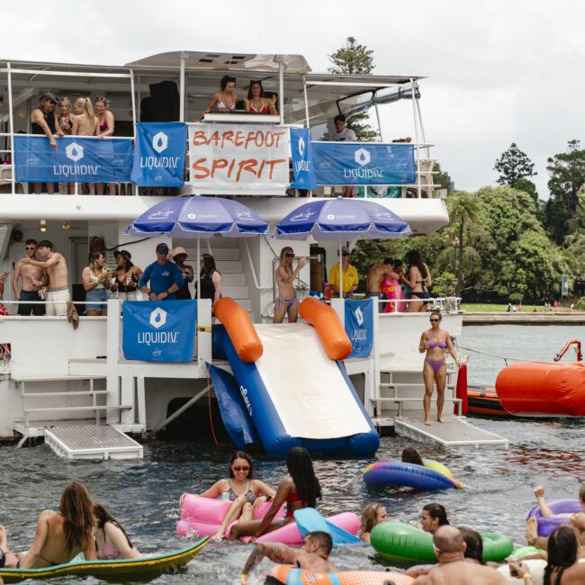 A lively boat party on a lake features people swimming with colorful inflatables. A slide extends from the boat, and several people are enjoying the water. A banner reading "Barefoot Spirit" hangs on the boat. Lush greenery is visible in the background.