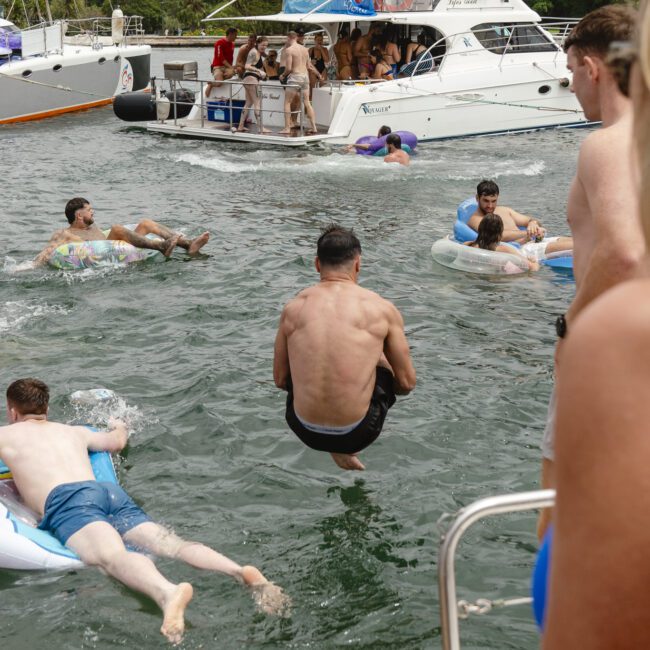 A man cannonballs into the water near a yacht filled with people, while others float on inflatable tubes. The scene is lively with greenery and city buildings in the background.