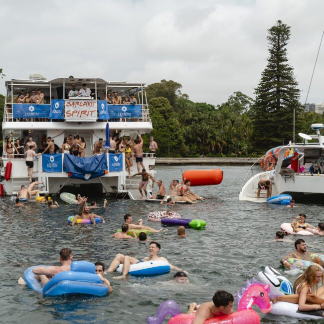 A lively scene on a lake with people enjoying a party. Two boats are docked nearby, one named "Barefoot Spirit." Many people are floating in the water on inflatables shaped like unicorns and other animals. The sky is overcast, and trees are in the background.